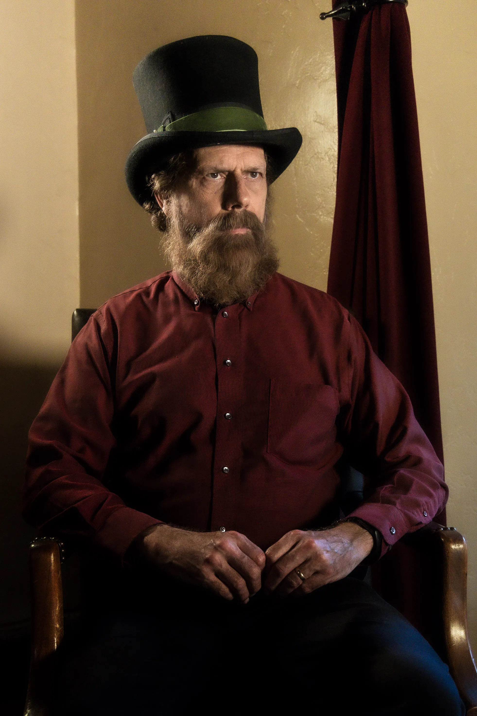 A costumed performer in a tall black hat and burgundy shirt sits with hands folded.