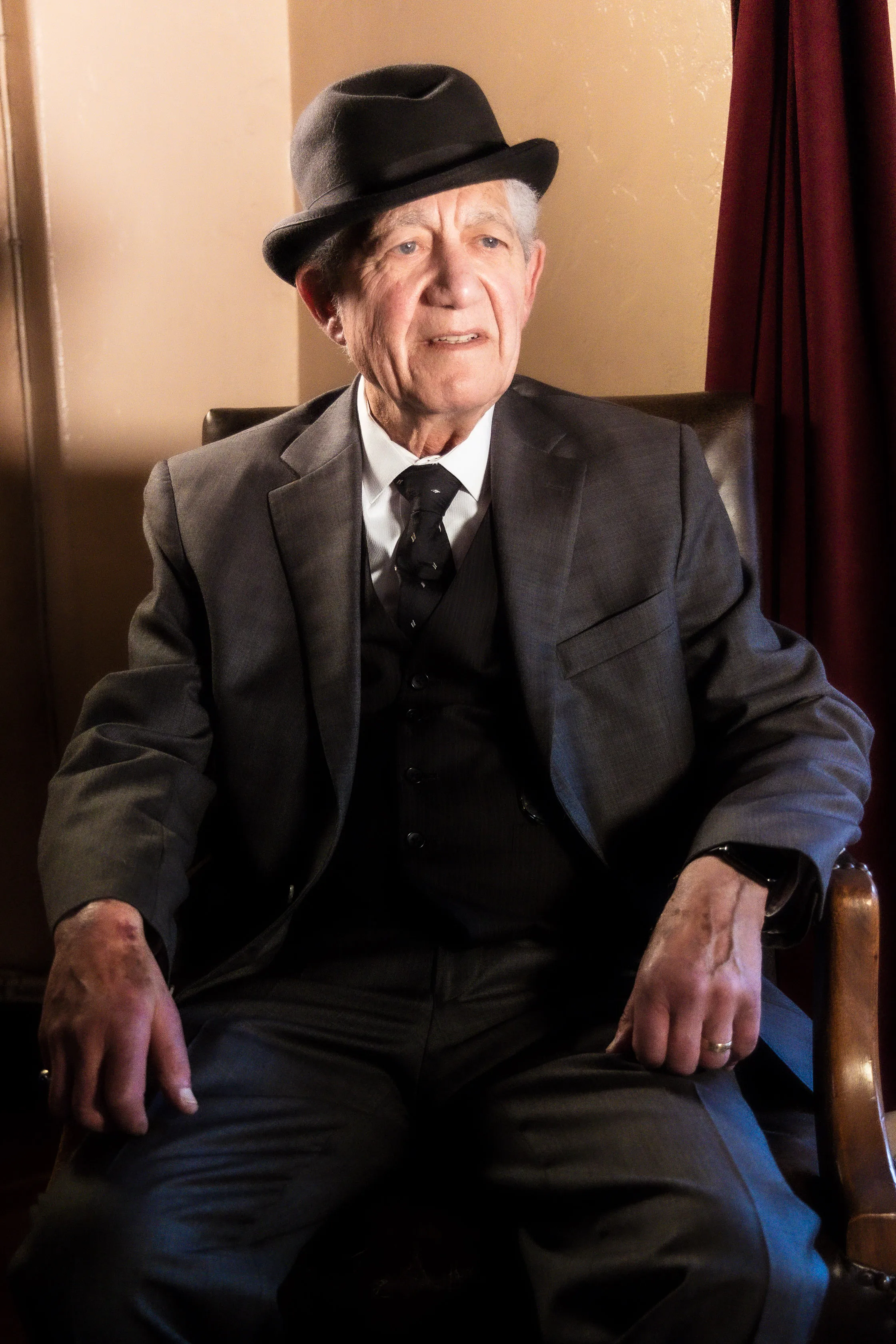 A costumed performer in a grey suit and black hat sits in an armchair.