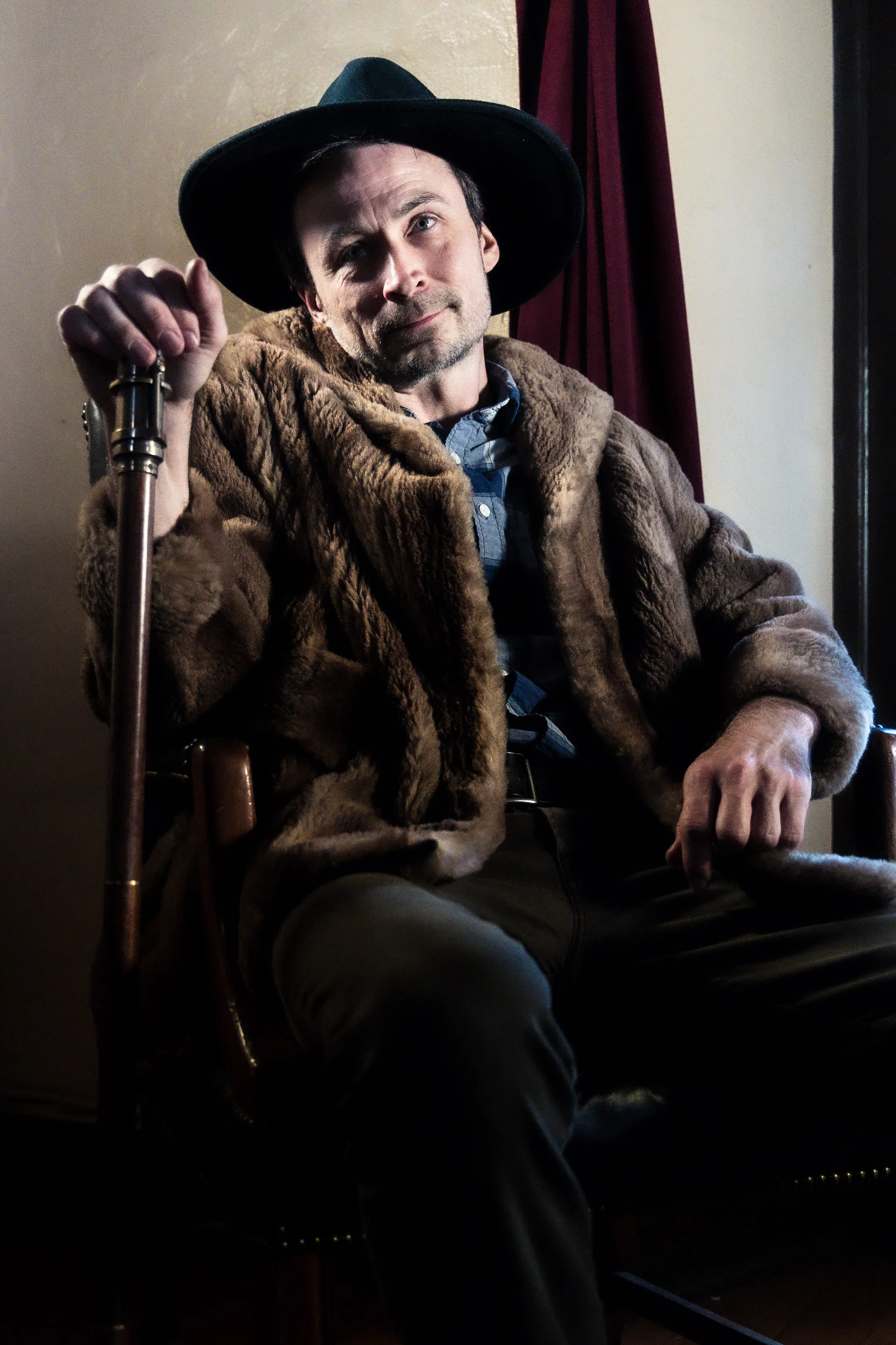 A performer in a fur coat and broad-brimmed hat sits holding a cane.