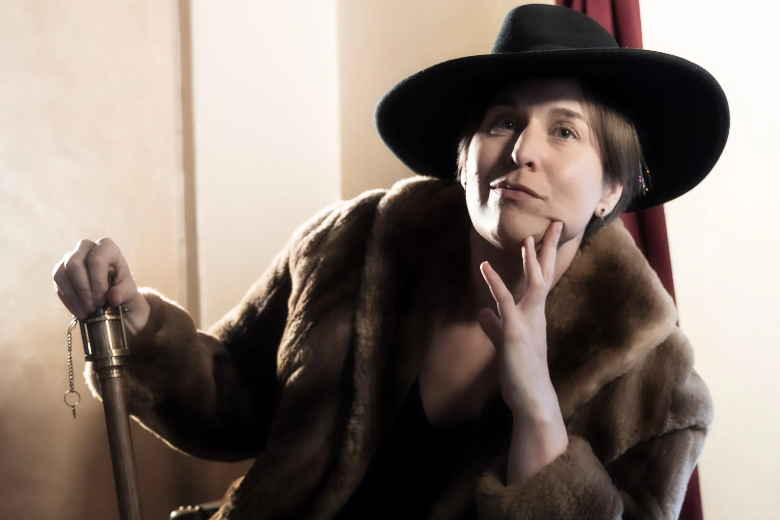 A seated performer in a fur coat rests one hand against their face and holds a cane.