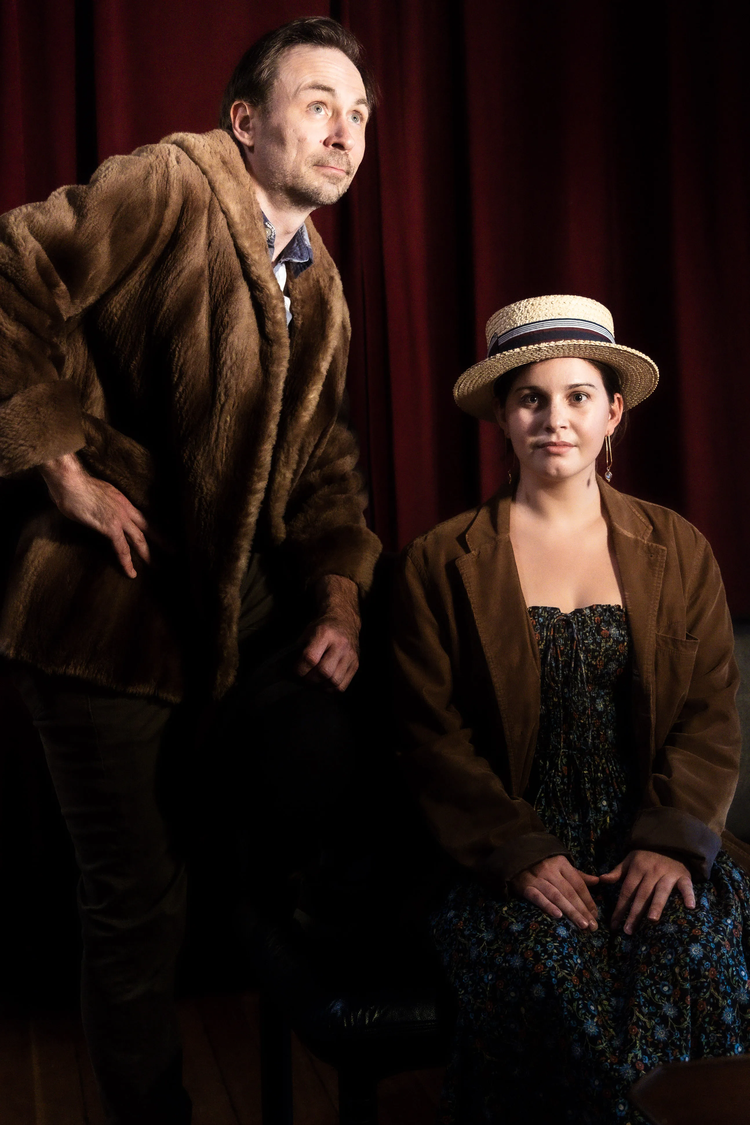 A performer in a fur coat stands beside a seated performer wearing a straw boater hat.