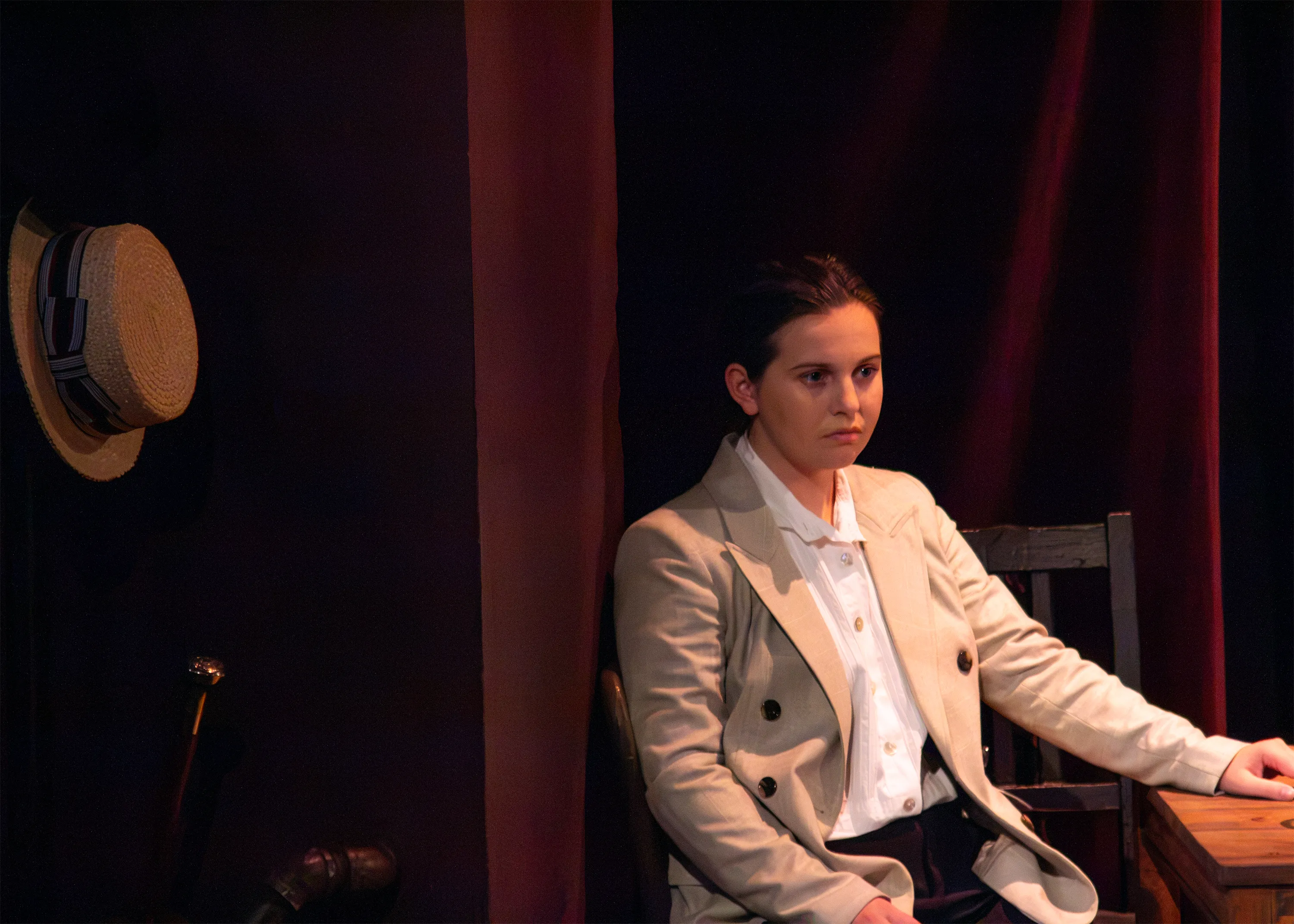 A performer in a pale suit sits beside a courtroom table, with a top hat hanging nearby.