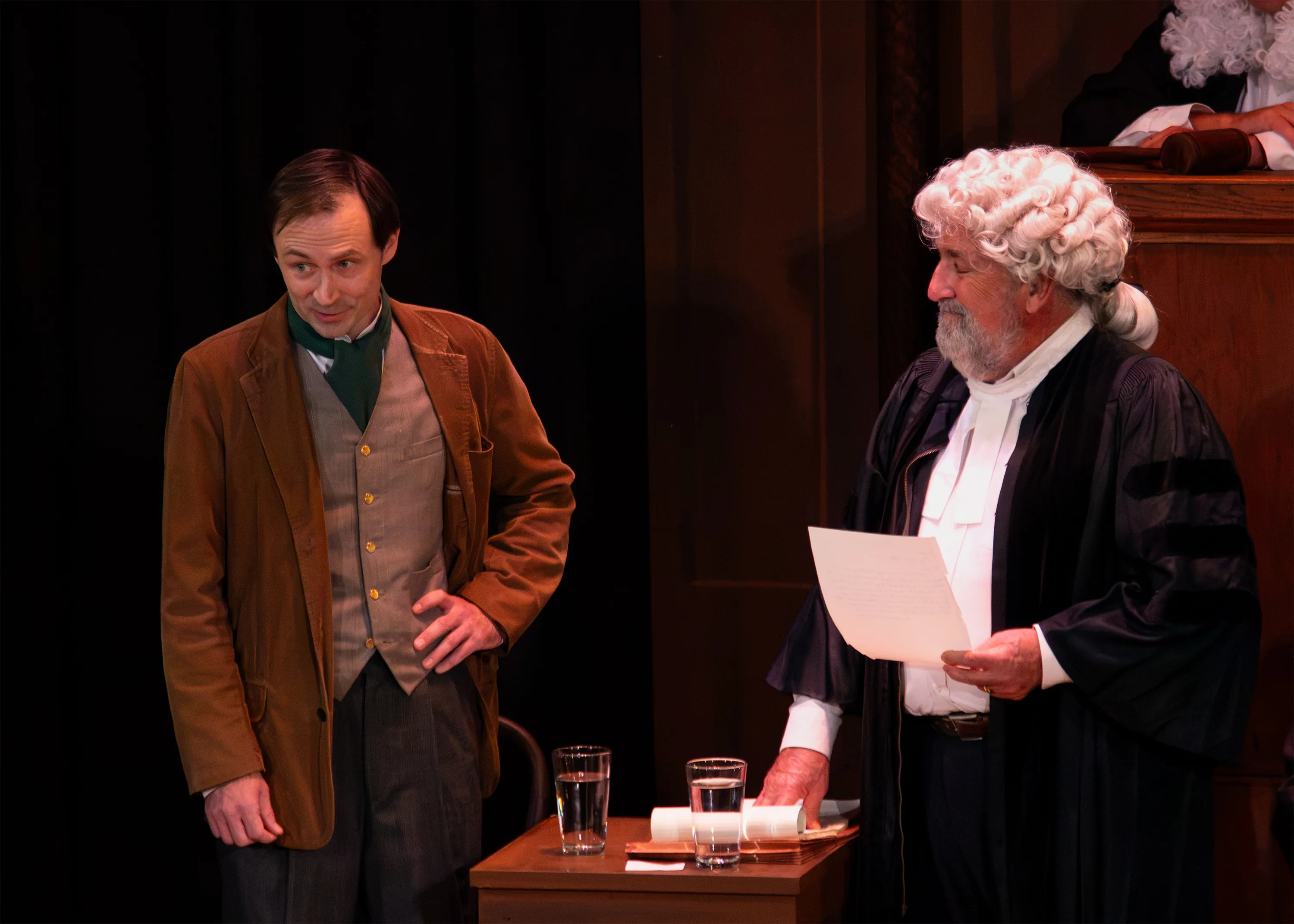A performer in a brown jacket listens to a barrister holding a sheet of paper.