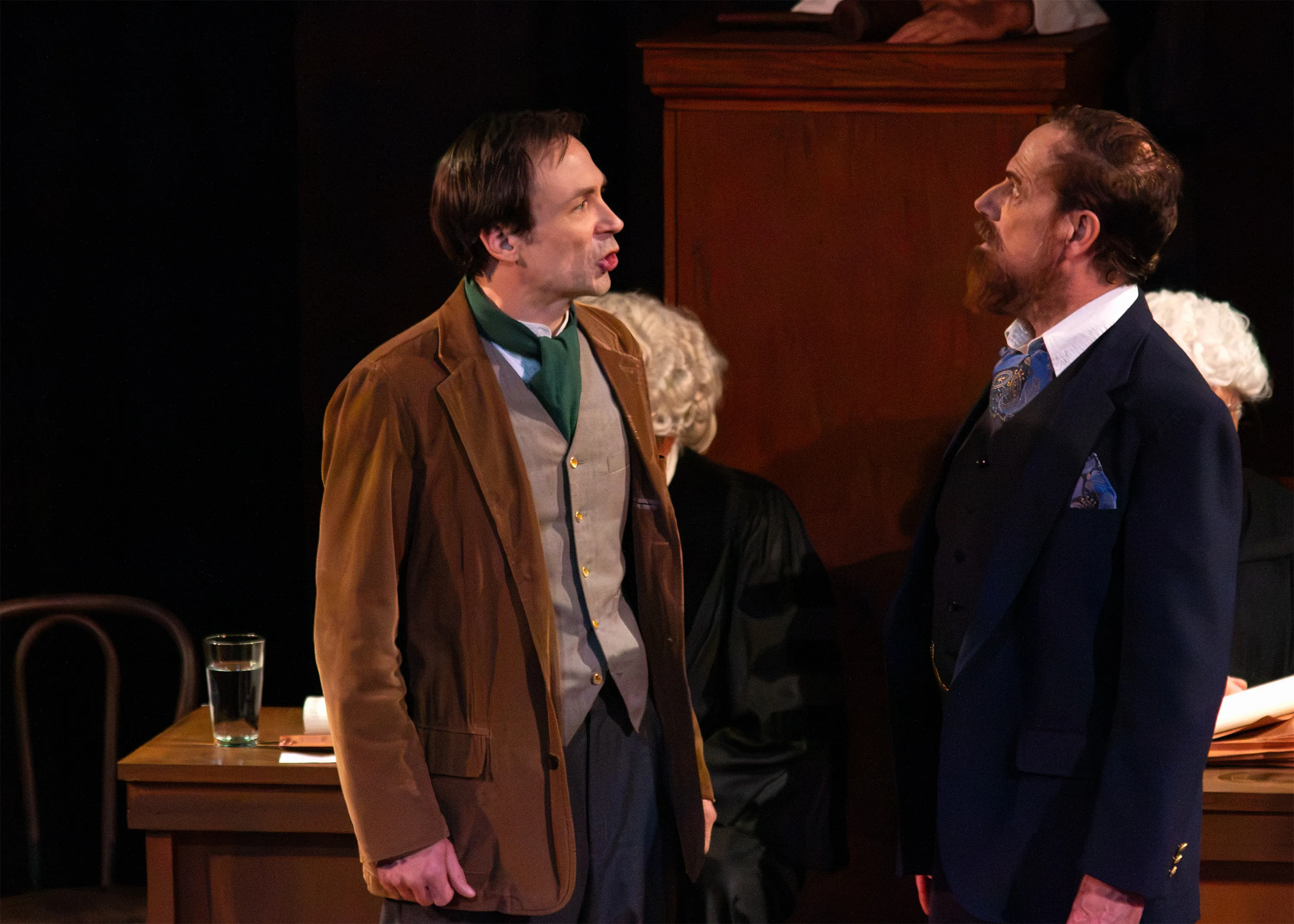 Two performers face one another in brown and navy jackets during a courtroom scene.