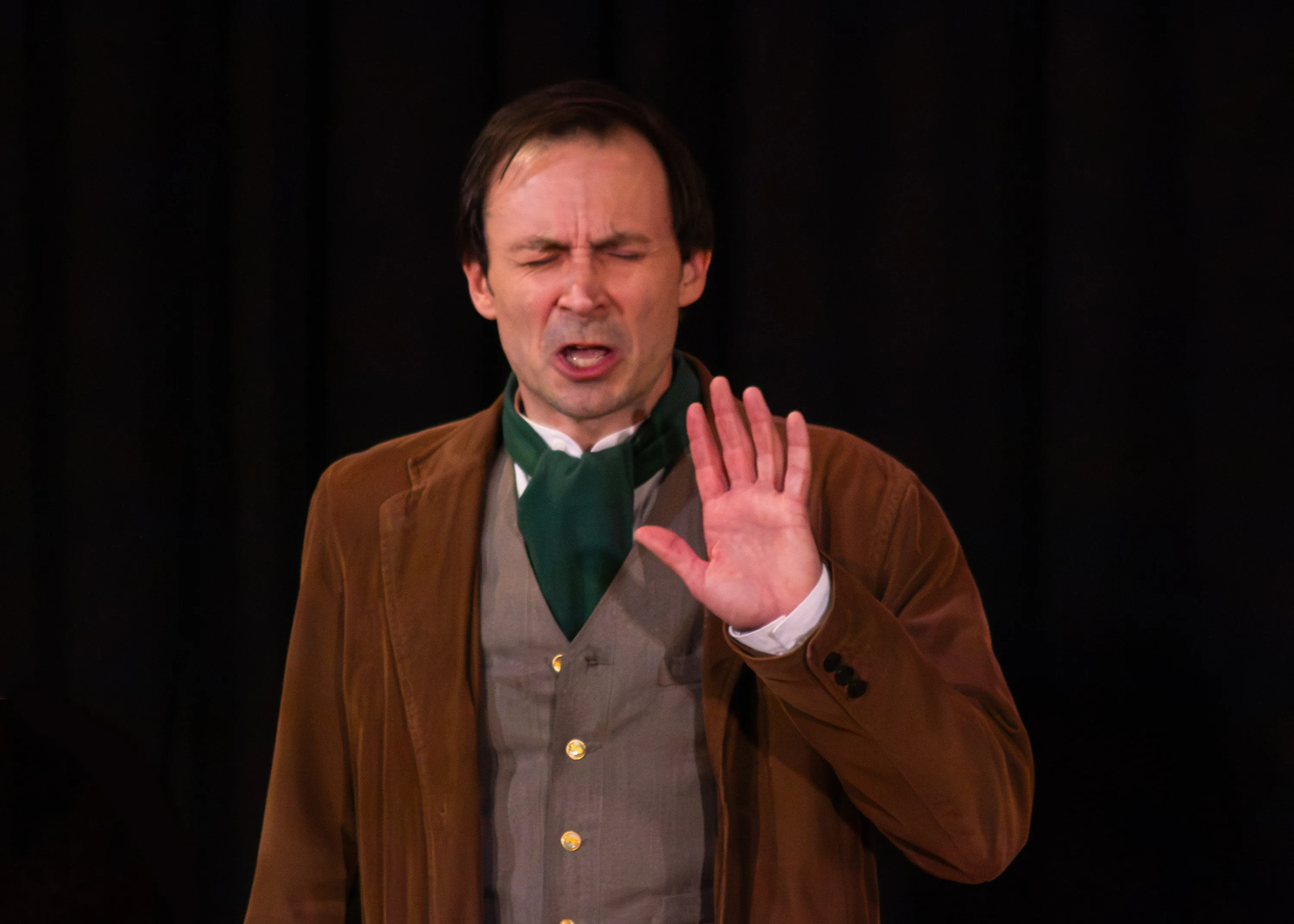 A performer in a brown jacket raises one hand while closing their eyes.
