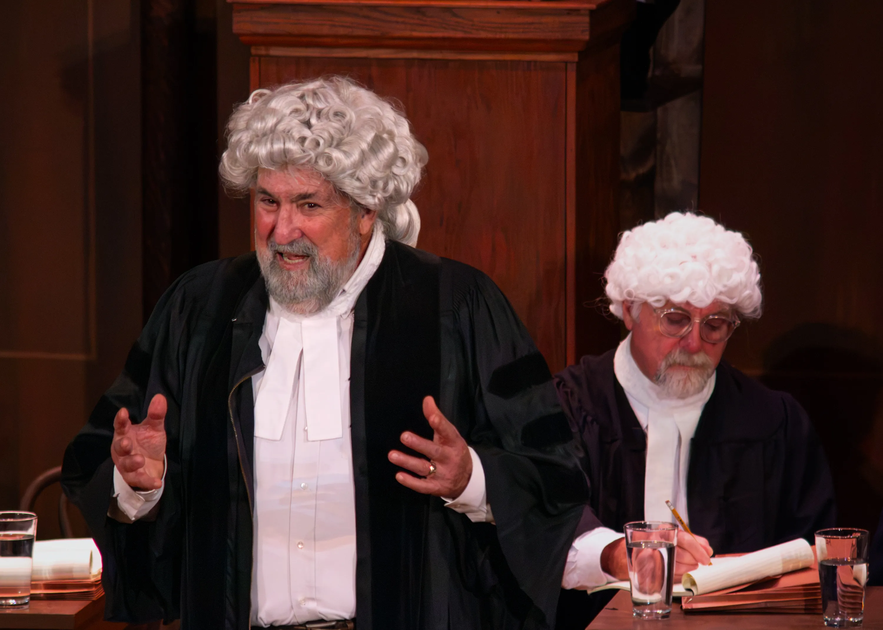 A barrister in a white wig gestures with both hands, with another court official seated behind.