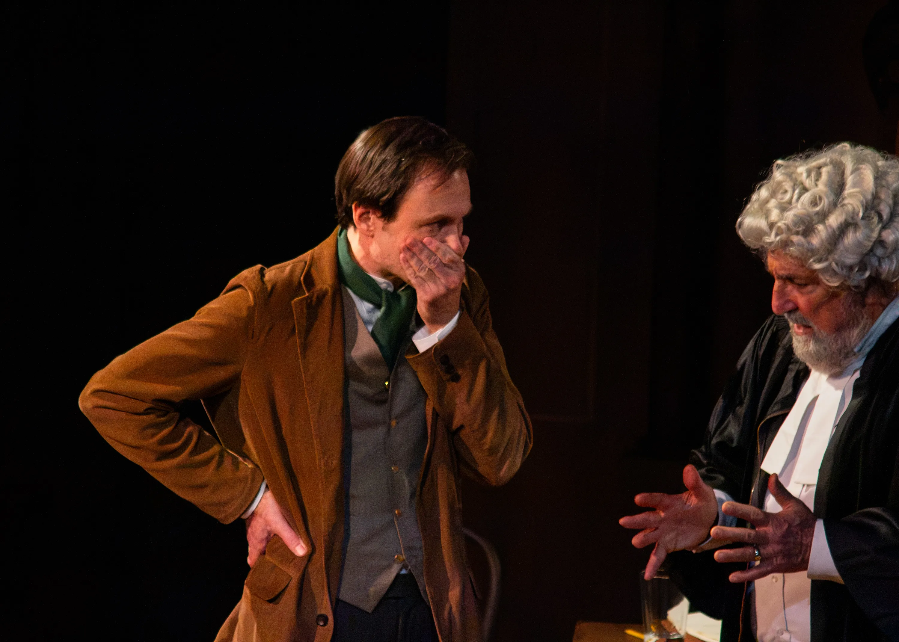 A performer in a brown jacket leans towards a barrister holding a paper.