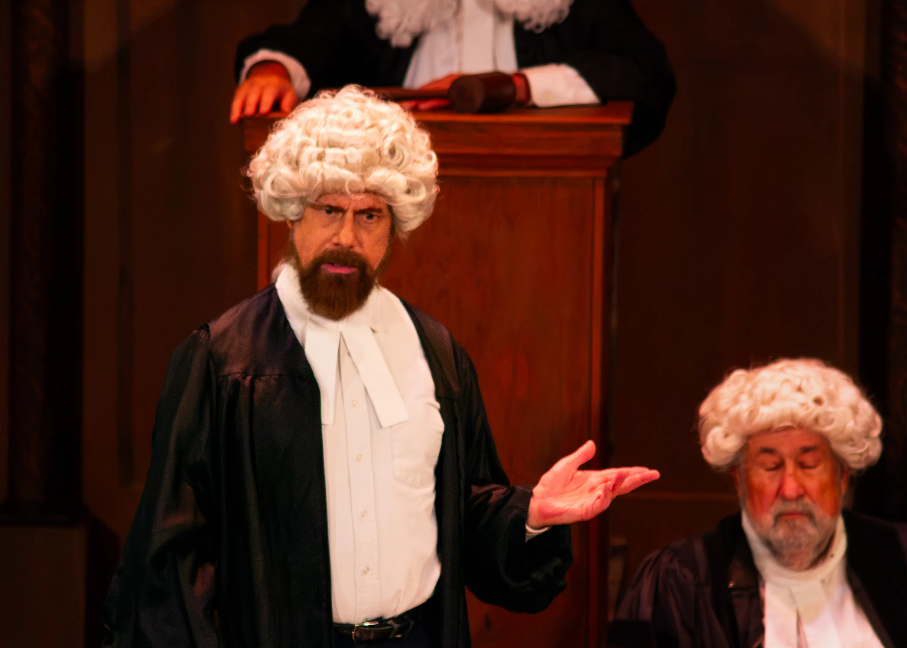 A barrister in a white wig extends one arm beside a seated court official.