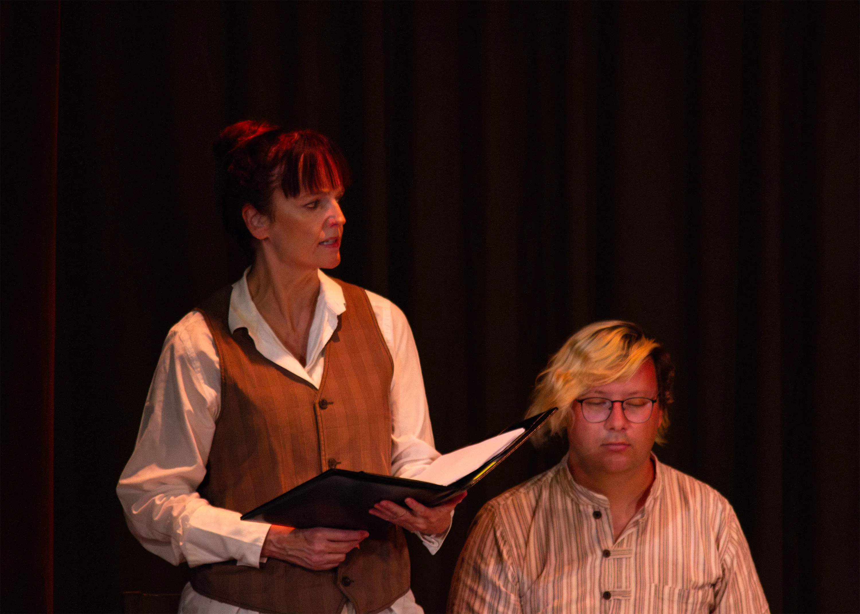 Two performers read from a binder, one wearing a waistcoat and the other a striped shirt.