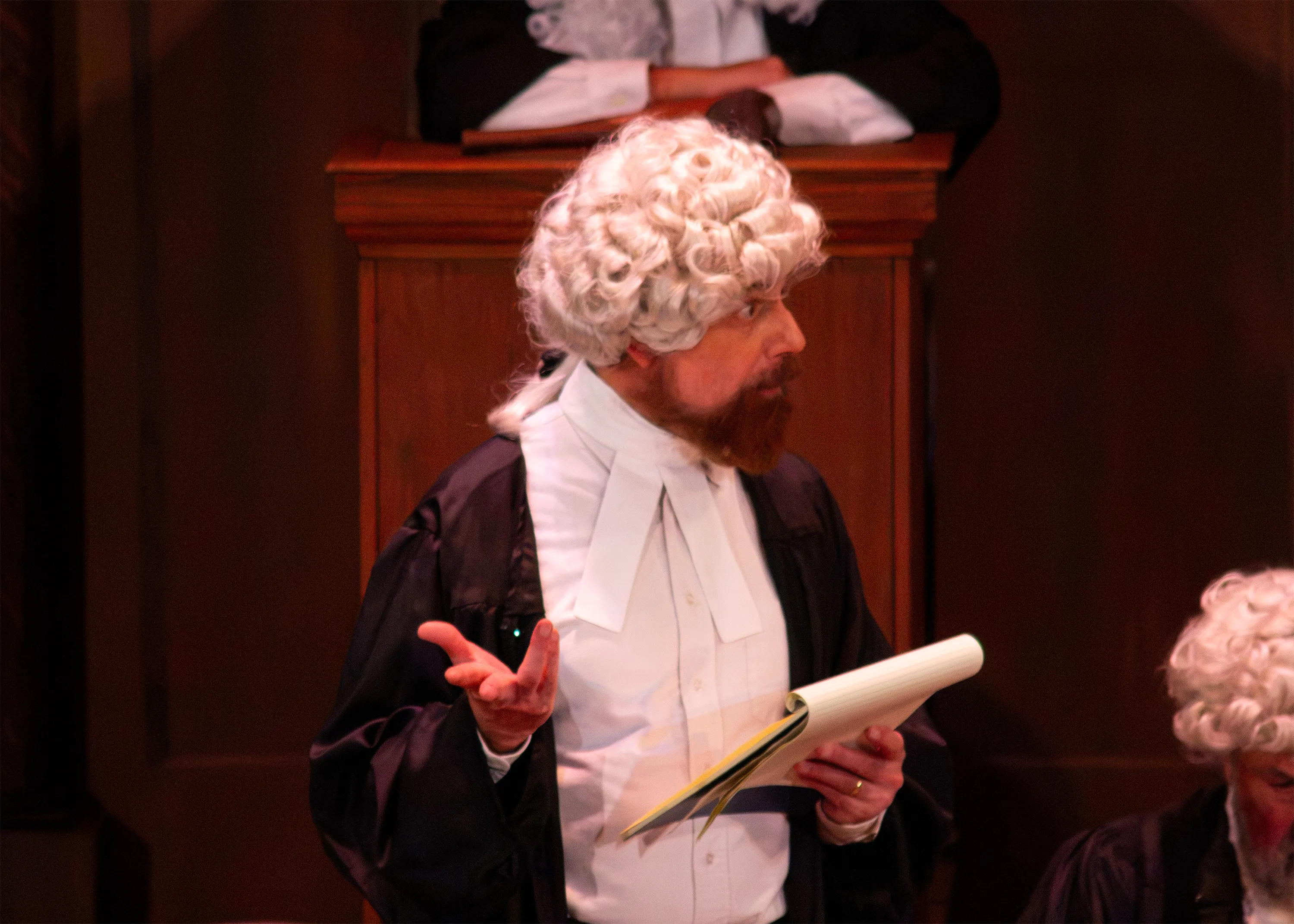 A barrister in a white wig reads from papers in front of the judge’s bench.