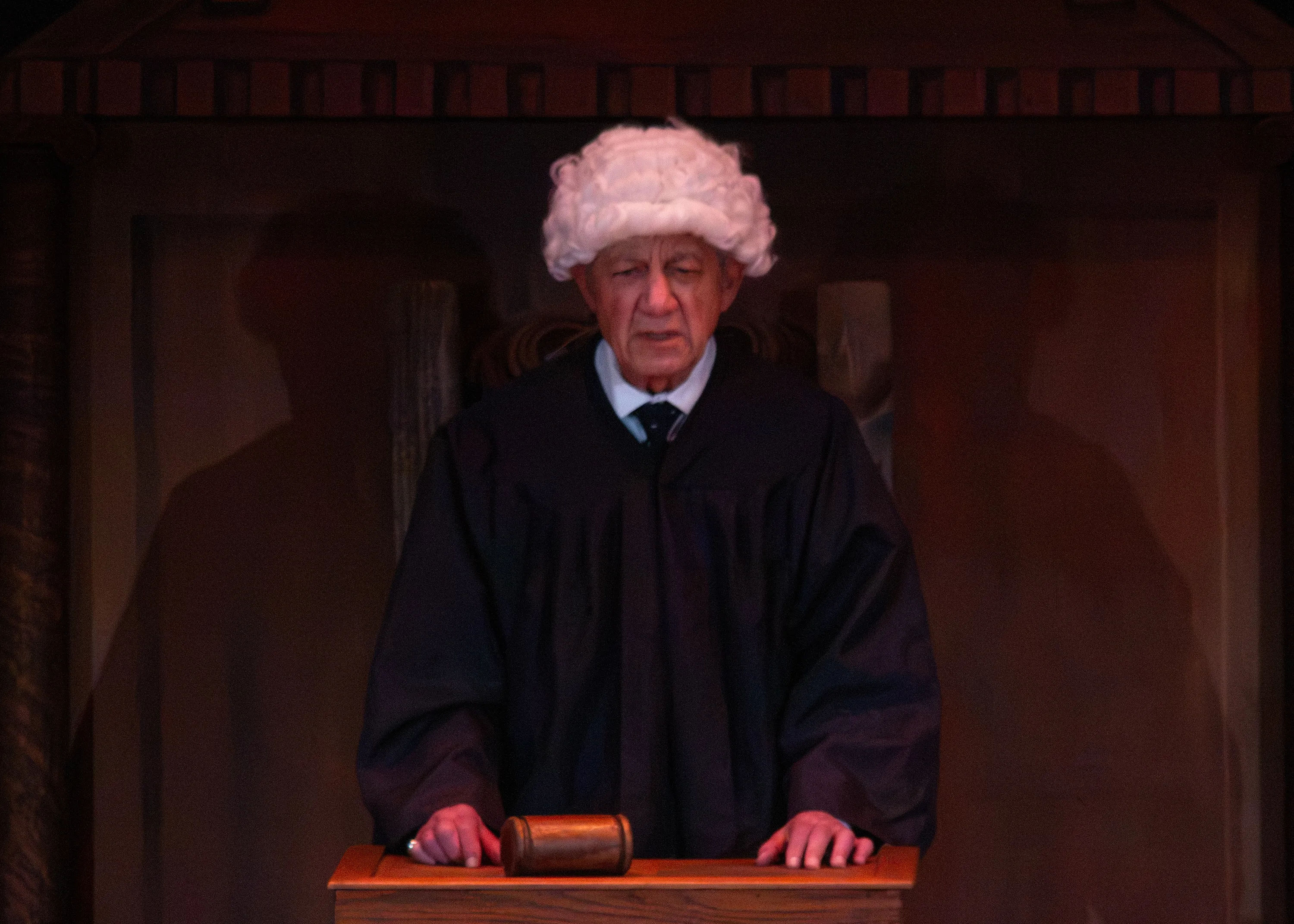 A judge in a black robe and white wig stands behind the wooden courtroom bench.